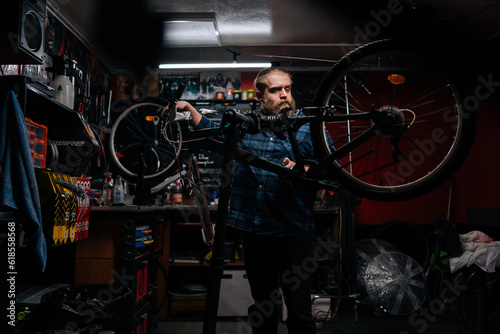 Wide shot portrait of repairman watching rear shifter of mountain bike, changing speeds using handlebar shift lever, working in bicycle repair shop with dark interior. Concept of maintenance of bike.