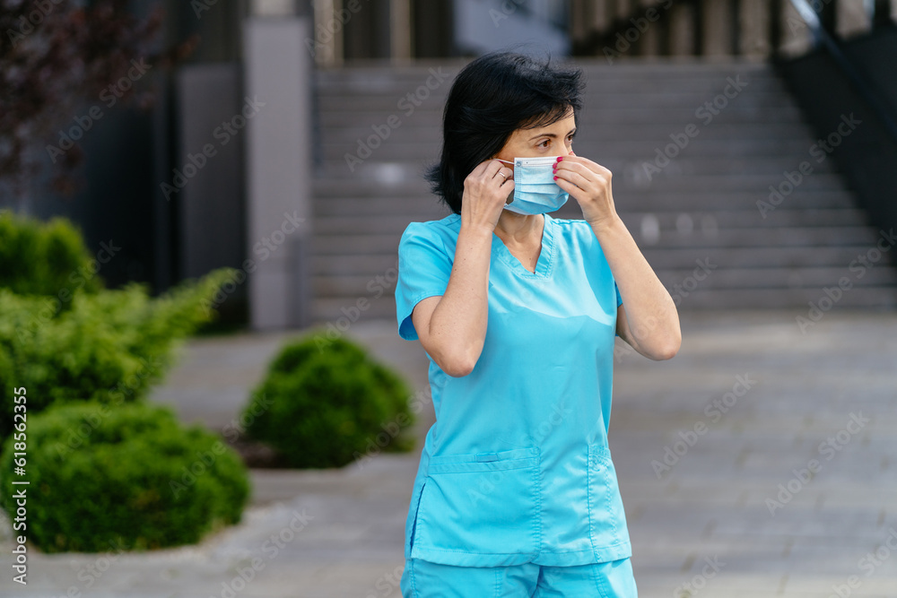 Female medical staff wearing medical protective mask while standing outdoor with modern hospital building on background. Profession, healthcare, medicine concept. Female medical staff wearing medical protective mask while standing outdoor with modern hospital building on background. Profession, healthcare, medicine concept.