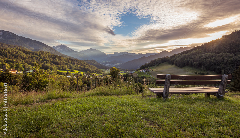 Wooden resting bank on the slope of a hill in the foreground, overlooking the valley below in Berchtesgaden, Germany. It is evening, during golden hour, clouds are covering the sun. Summer, day.