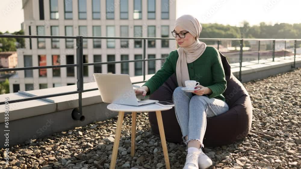 Charming muslim lady in headscarf holding cup of hot drink while scrolling webpages on portable computer outdoors. Delighted woman taking pleasure of online shopping during coffee break on rooftop. Charming muslim lady in headscarf holding cup of hot drink while scrolling webpages on portable computer outdoors. Delighted woman taking pleasure of online shopping during coffee break on rooftop.