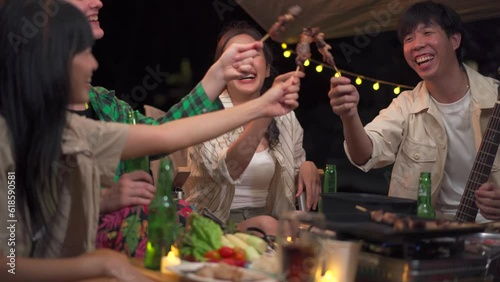 Group of diverse ethnicity young people enjoy eating and grilling bar-b-q together while camping in forest at night.