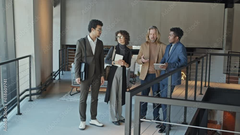 Full arc shot of group of diverse business people with bags standing on second floor passway in spacious office or hotel lobby, looking at tablet computer, discussing something while waiting for taxi Full arc shot of group of diverse business people with bags standing on second floor passway in spacious office or hotel lobby, looking at tablet computer, discussing something while waiting for taxi
