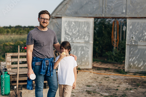 Father and son standing in front of a greenhouse and eating an apple from their garden.