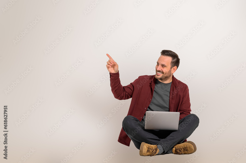 Cheerful marketing manager pointing at copy space for advertisement while sitting with legs crossed on background. Smiling young businessman showing empty copy space and working over laptop Cheerful marketing manager pointing at copy space for advertisement while sitting with legs crossed on background. Smiling young businessman showing empty copy space and working over laptop