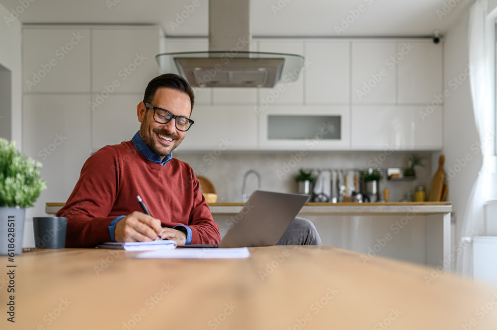 Smiling male entrepreneur writing project ideas in note pad and working on laptop at home office