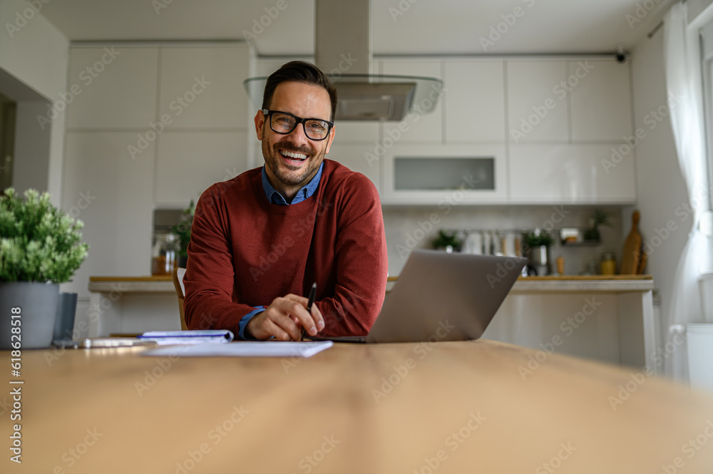 Portrait of smiling handsome businessman with glasses on working over laptop at desk in home office