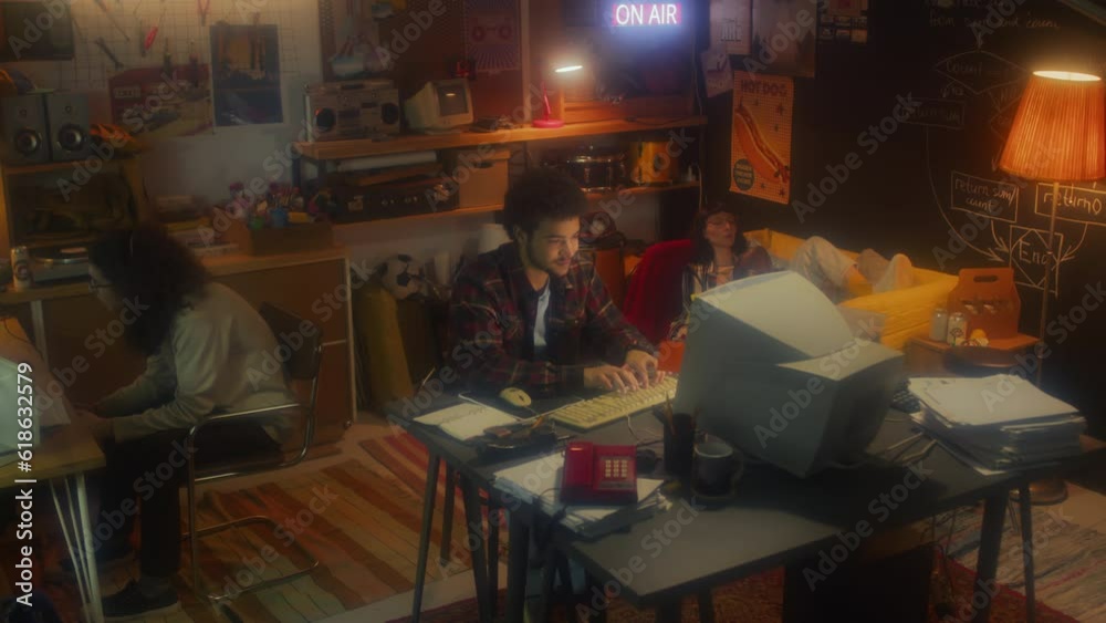 Wide shot of young Middle Eastern man working on desktop computer while his colleagues playing video game and relaxing Wide shot of young Middle Eastern man working on desktop computer while his colleagues playing video game and relaxing