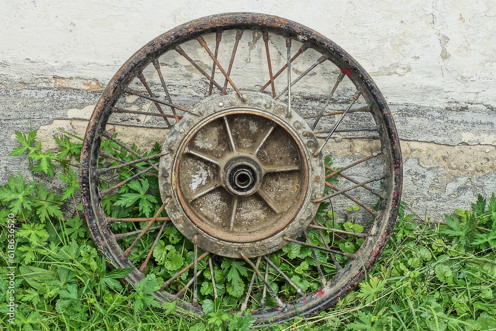 an iron rusty disc with thick iron rusty spokes from an old wheel of a retro motorcycle stands outdoors on green grass near a white wall