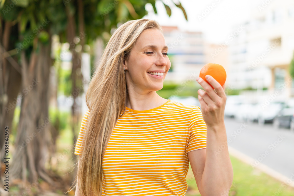 Young blonde woman holding an orange at outdoors with happy expression Young blonde woman holding an orange at outdoors with happy expression