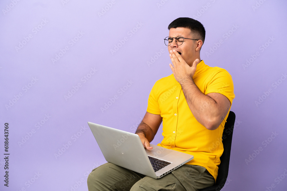 Young man sitting on a chair with laptop yawning and covering wide open mouth with hand Young man sitting on a chair with laptop yawning and covering wide open mouth with hand