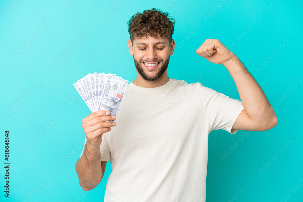 Young caucasian man taking a lot of money isolated on blue background doing strong gesture Young caucasian man taking a lot of money isolated on blue background doing strong gesture