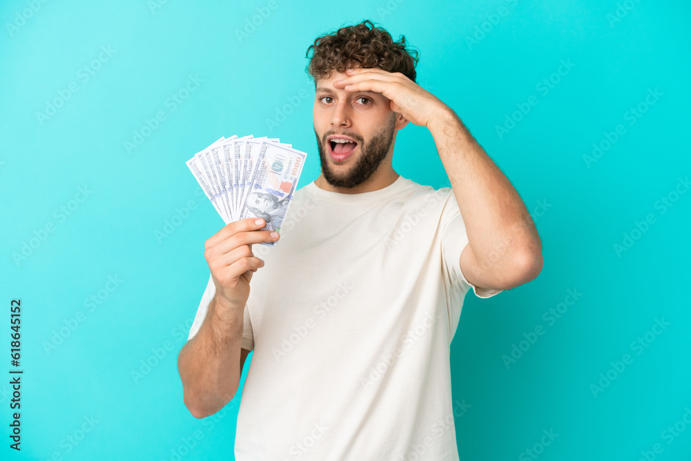 Young caucasian man taking a lot of money isolated on blue background doing surprise gesture while looking to the side Young caucasian man taking a lot of money isolated on blue background doing surprise gesture while looking to the side