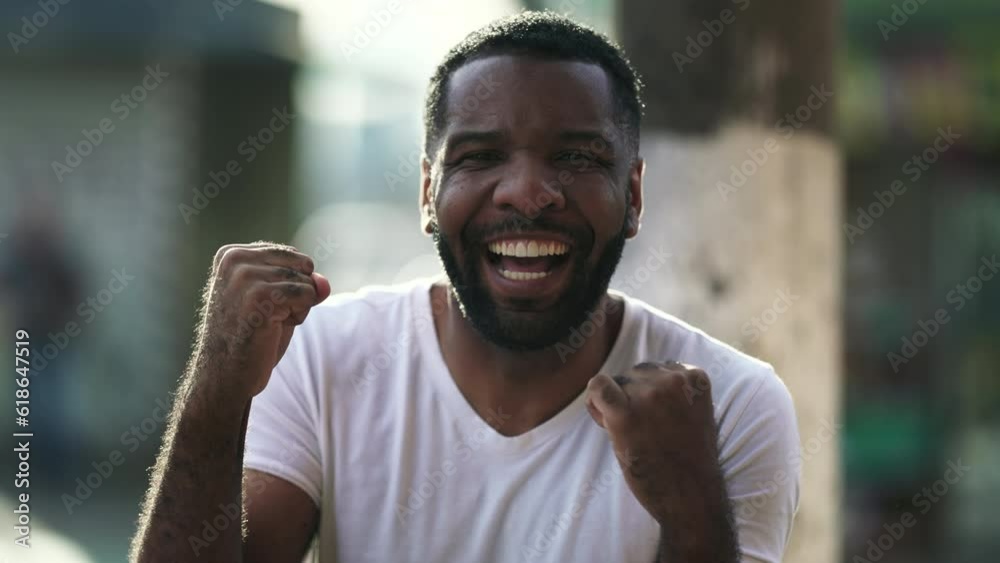 One Excited Black Man Celebrating Success, Raising Clenched Fists in Air with Happiness standing in urban environment in daylight One Excited Black Man Celebrating Success, Raising Clenched Fists in Air with Happiness standing in urban environment in daylight