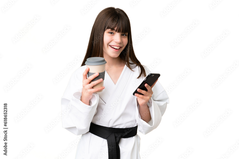 Little Caucasian girl doing karate over isolated background holding coffee to take away and a mobile Little Caucasian girl doing karate over isolated background holding coffee to take away and a mobile