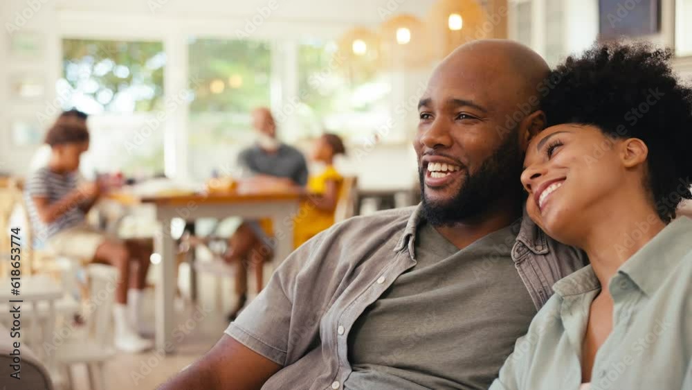 Couple at home on sofa streaming show or movie to TV with multi-generation family in background - shot in slow motion Couple at home on sofa streaming show or movie to TV with multi-generation family in background - shot in slow motion
