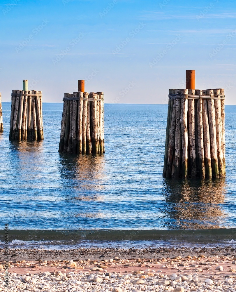 Posts in a marina surrounded by wood in calm waters of a bay with a rocky beach, blue sky behind. Coastal summer mood. Posts in a marina surrounded by wood in calm waters of a bay with a rocky beach, blue sky behind. Coastal summer mood.