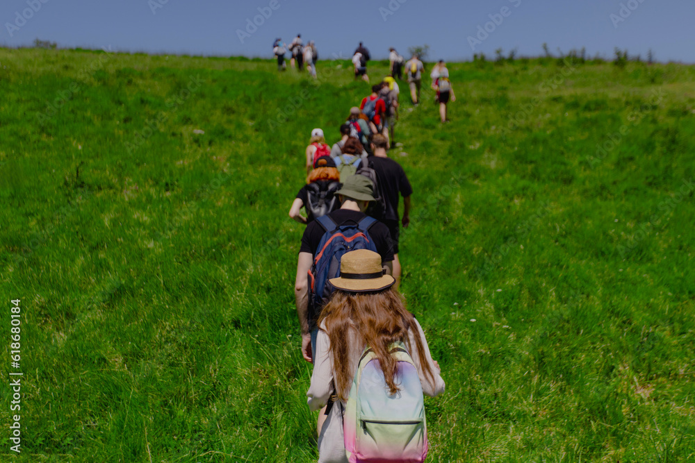 Group of hikers with backpacks climbing hill with green meadow on summer sunny day