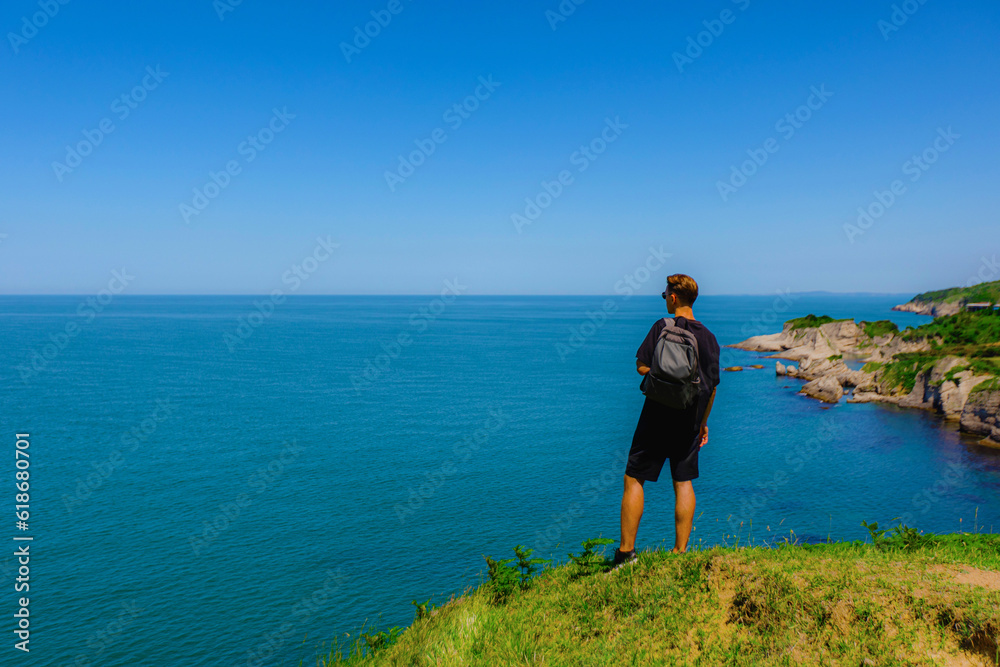 Tourist in black with backpack on rocks by sea on summer sunny day