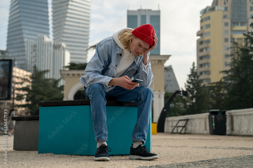Frustrated pensive hipster guy looks at phone while sitting on bench against backdrop of modern cityscape. Confused man read bad news and sad conversation. Guy was dumped by girl on phone