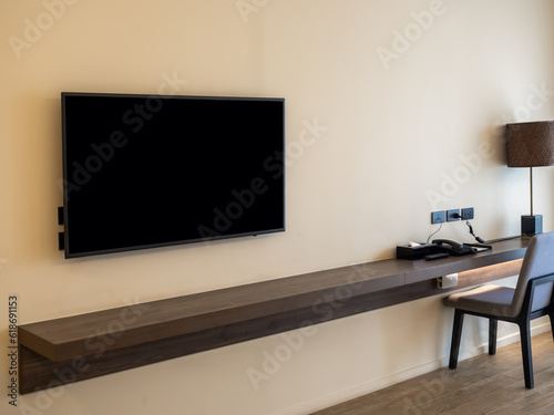 Mockup blank space on smart TV black screen over the empty long brown walnut wooden table shelf with chair at working area on white wall background with yellow warm lighting in the hotel bedroom.