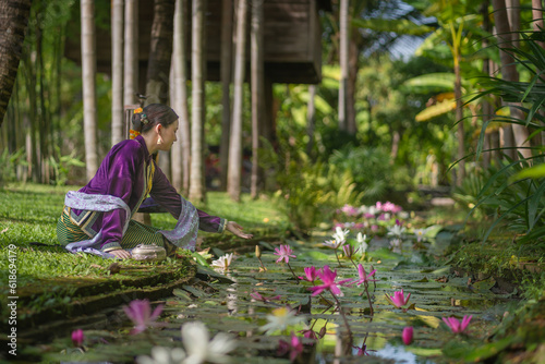 A pretty young Asian woman wearing an elegant luxurious traditional Chiang Mai dress costume sits on a natural green lawn by a stream with a beautiful pink lotus flower nearby. © pomphotothailand