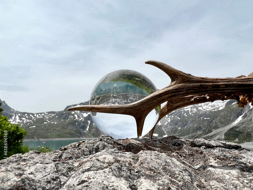 Fototapeta premium Roebuck antlers next to Lensball, crystal ball, with reflections of Lake Lunersee (Lünersee, Montafon, Vorarlberg). In the background the famous high mountains of austria and switzerland (Rätikon).