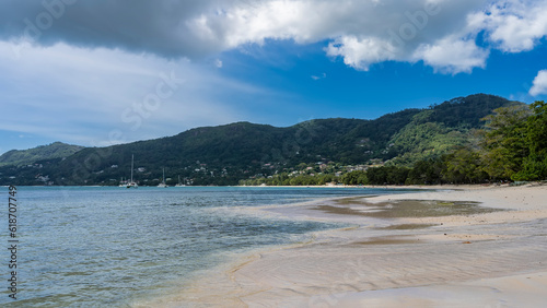 Fototapeta Naklejka Na Ścianę i Meble -  The waves of the turquoise ocean spread over the beach. Wet sand glistens. Yachts in the distance. A green hill against a background of blue sky and clouds. Seychelles. Mahe. Beau Vallon.