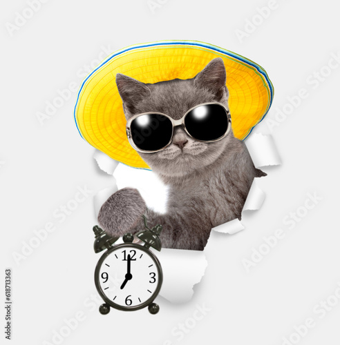 Cute kitten wearing sunglasses and summer hat looks through the hole in white paper and shows alarm clock