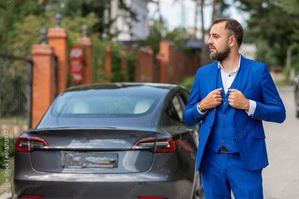 Caucasian bearded man in a blue suit stands near a black car in the countryside in summer.