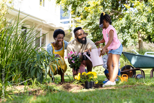 African american cheerful parents with children planting and watering flowers on field in backyard