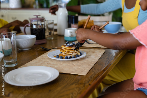 Midsection of african american girl eating pancakes with fork and table knife at dining table