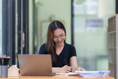 A beautiful young Asian businesswoman is saving and checking work and documents with her laptop computer at her desk with a bright smile on her face.