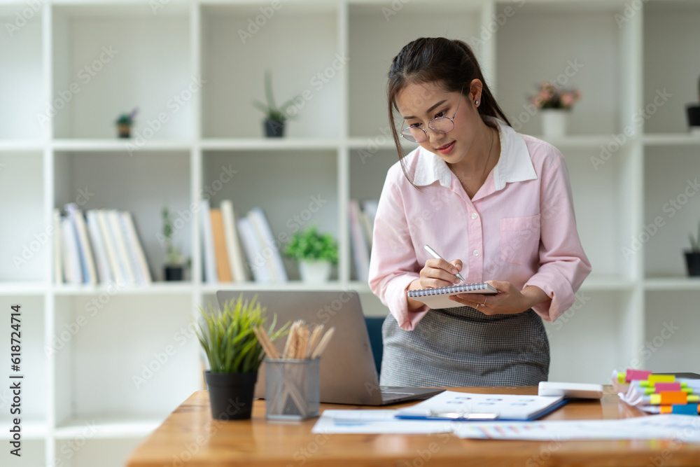 Office worker or businesswoman, attractive Asian woman in eyeglasses researching Analyze information about the business plan. take notes in a book Notebook while using a laptop computer at her desk. Office worker or businesswoman, attractive Asian woman in eyeglasses researching Analyze information about the business plan. take notes in a book Notebook while using a laptop computer at her desk.