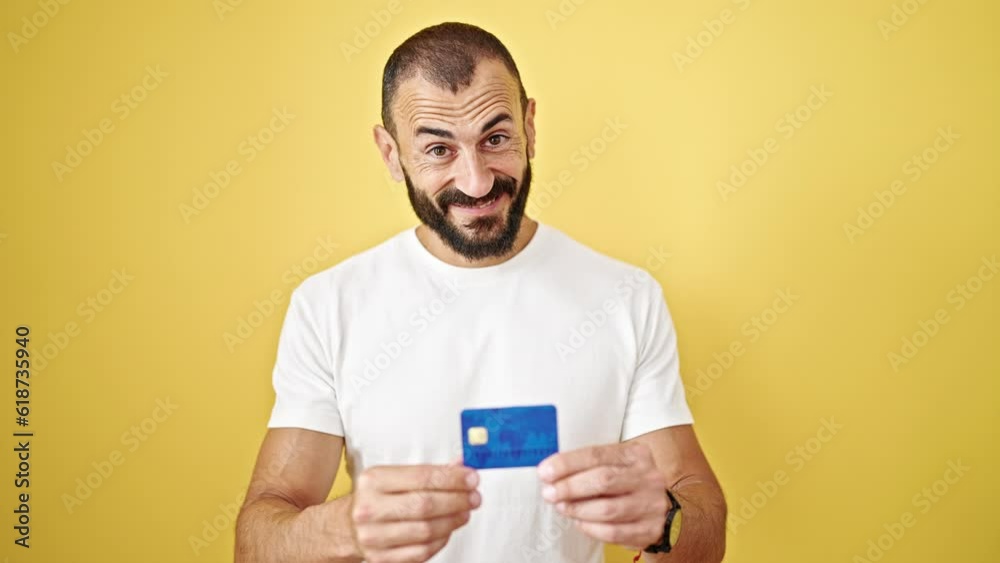 Young hispanic man pointing to credit card with thumb up gesture over isolated yellow background Young hispanic man pointing to credit card with thumb up gesture over isolated yellow background
