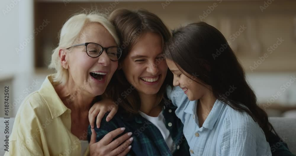 Laughing multi generational women cuddling, tickling each other, having fun seated on sofa at home, close up. Playful retired 60s grandma spend carefree leisure with grandkid and daughter. Family ties