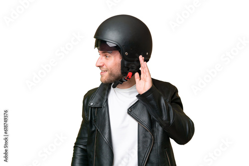 Young caucasian man with a motorcycle helmet isolated on green chroma background listening to something by putting hand on the ear