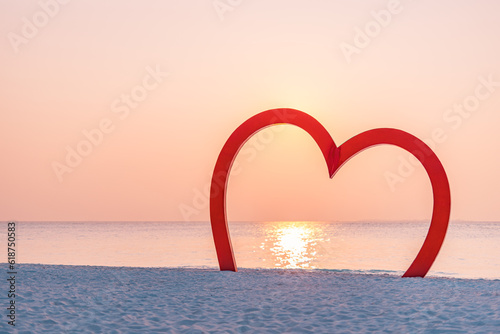 Golden sunset sunlight over wooden red heart in sandy beach. Romance love travel concept. Sea sky view, couple honeymoon anniversary beach decoration. Idyllic romantic destination wedding background