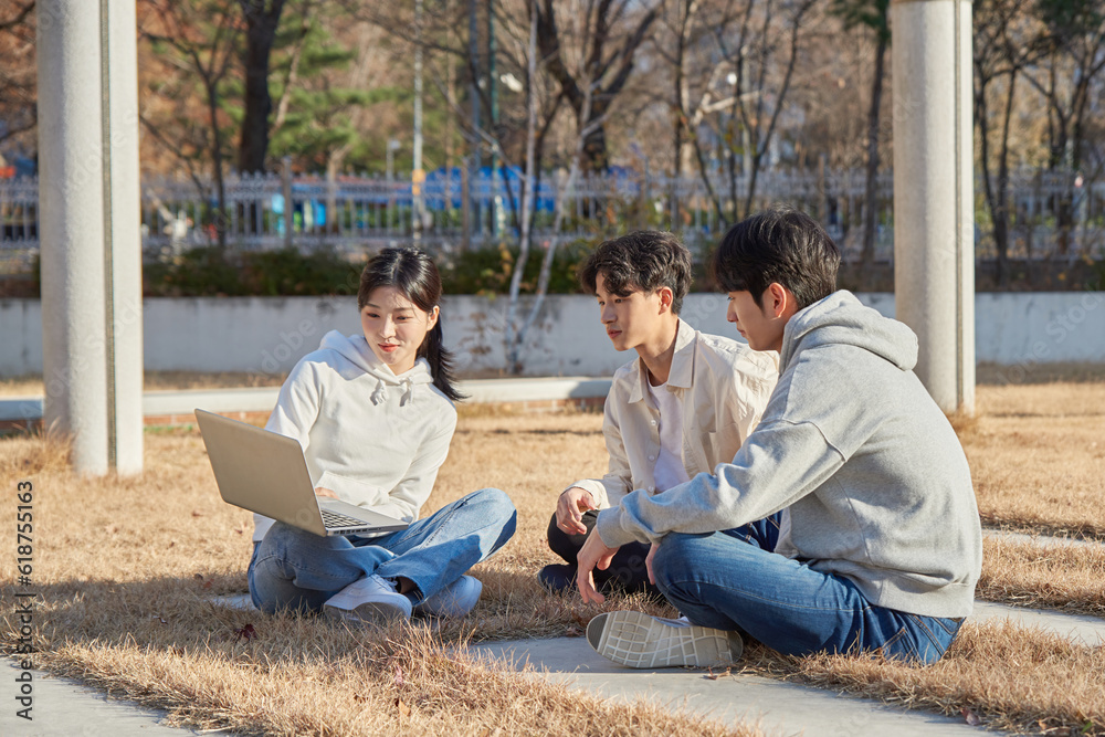 Three young college student male and female models sitting on the lawn at the university in autumn in Asian Korea, watching a laptop, listening to a lecture, discussing or talking