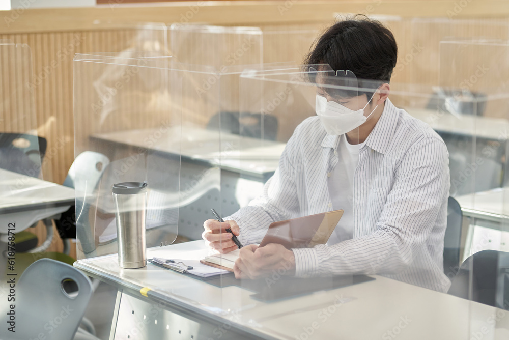A young male Asian college student is learning or listening to a lesson while her face is outside a protective shield on the desk in a university classroom in Korea during the pandemic. A young male Asian college student is learning or listening to a lesson while her face is outside a protective shield on the desk in a university classroom in Korea during the pandemic.