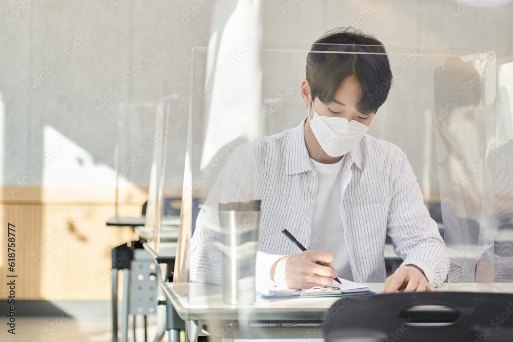 A young male Asian college student is learning or listening to a lesson while her face is outside a protective shield on the desk in a university classroom in Korea during the pandemic. A young male Asian college student is learning or listening to a lesson while her face is outside a protective shield on the desk in a university classroom in Korea during the pandemic.