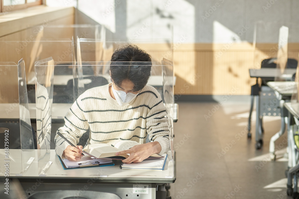 A young male Asian college student is learning or listening to a lesson while her face is outside a protective shield on the desk in a university classroom in Korea during the pandemic. A young male Asian college student is learning or listening to a lesson while her face is outside a protective shield on the desk in a university classroom in Korea during the pandemic.