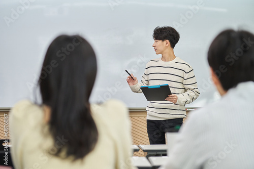 An Asian young man is standing in front of a lecture hall at a university in South Korea, giving a presentation or lecture. In front of him are male and female students. 