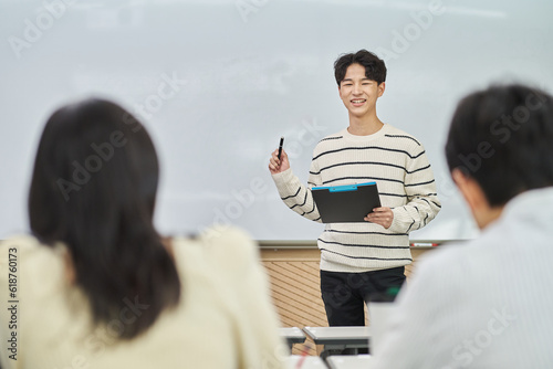 An Asian young man is standing in front of a lecture hall at a university in South Korea, giving a presentation or lecture. In front of him are male and female students. 