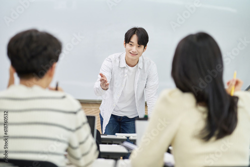 An Asian young man is standing in front of a lecture hall at a university in South Korea, giving a presentation or lecture. In front of him are male and female students.  © TEAM PRE-LIGHT