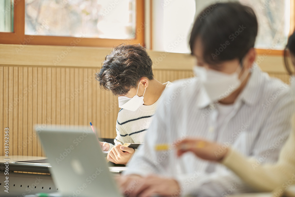 In a higher education classroom in South Korea, young university students wearing masks are listening to a lecture, studying, and talking. A woman and a man are in the background In a higher education classroom in South Korea, young university students wearing masks are listening to a lecture, studying, and talking. A woman and a man are in the background