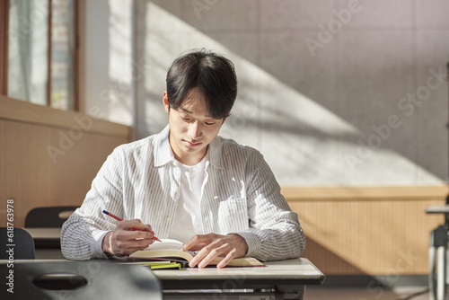 A young college student is sitting at a desk in a university classroom in South Korea, Asia. He is either reading a book or looking out of the window