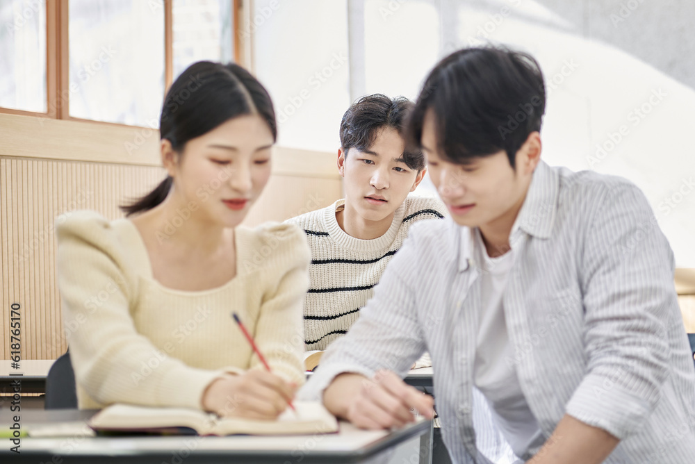Three young male and female college students models sitting or standing at desks in a university classroom in South Korea, Asia, talking or having a discussion Three young male and female college students models sitting or standing at desks in a university classroom in South Korea, Asia, talking or having a discussion