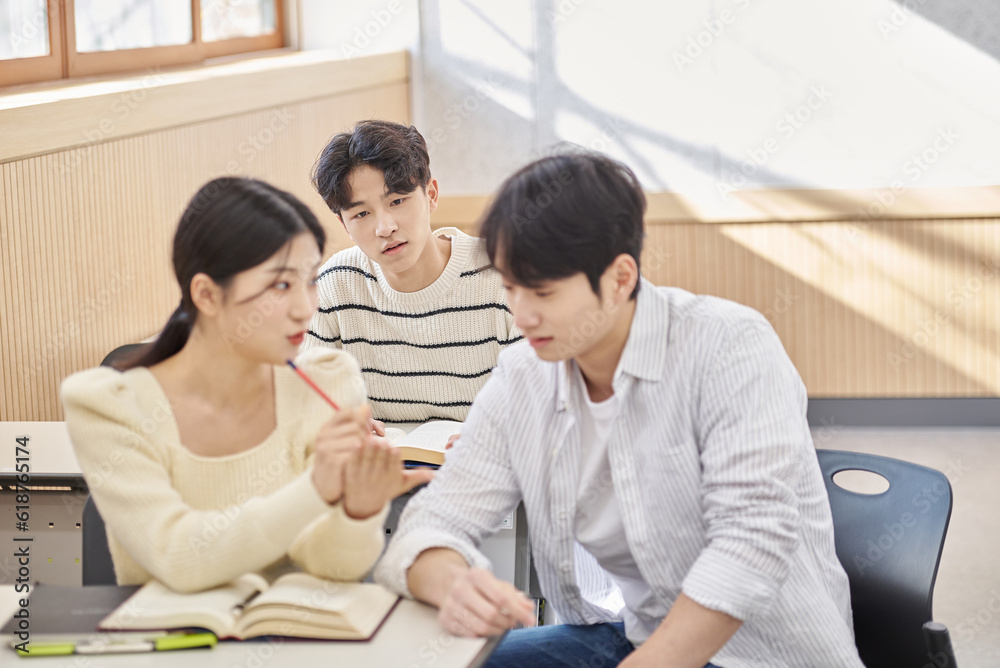 Three young male and female college students models sitting or standing at desks in a university classroom in South Korea, Asia, talking or having a discussion Three young male and female college students models sitting or standing at desks in a university classroom in South Korea, Asia, talking or having a discussion