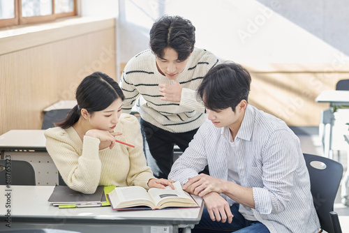 Three young male and female college students models sitting or standing at desks in a university classroom in South Korea, Asia, talking or having a discussion