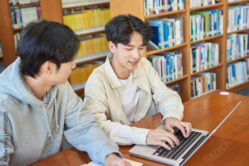 A male student sits at a desk in a university library in South Korea, Asia, looking at a laptop computer with bookshelves of books in the background.